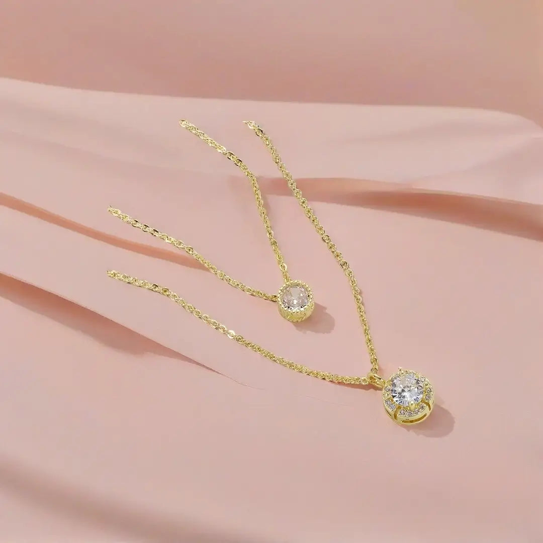 Elegant gold layered necklace featuring two pendants: a small solitaire and a large round halo pendant with pave-set crystals, laid on a blush pink fabric background.